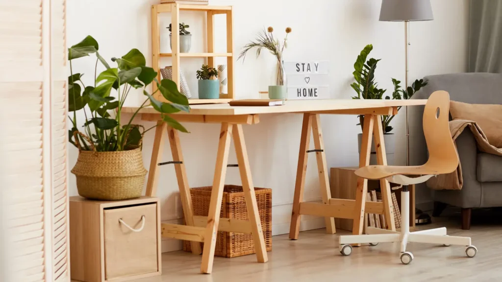 DIY Home Office Ideas on a Budget - A bright, budget-friendly home office setup in a small room featuring a DIY desk made from a wooden countertop on trestle legs, a thrifted chair with a cushion, floating shelves with plants, and a simple desk lamp