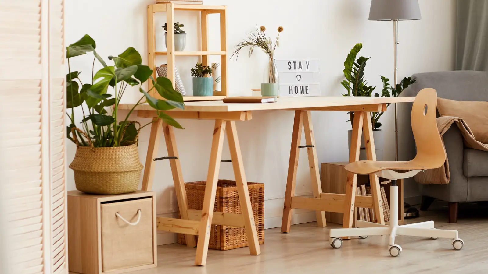 DIY Home Office Ideas on a Budget - A bright, budget-friendly home office setup in a small room featuring a DIY desk made from a wooden countertop on trestle legs, a thrifted chair with a cushion, floating shelves with plants, and a simple desk lamp