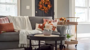 A cozy fall living room with burnt orange and forest green throw pillows, a chunky wool blanket, dried grasses in a ceramic vase, and warm amber candlelight.