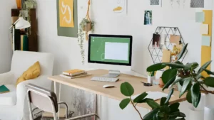 A small apartment corner desk setup for women with a floating shelf above, a compact plant, a slim lamp, and minimal surface decor in a bright, tight space
