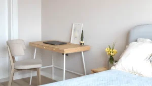 Small feminine home office in a bedroom corner with a slim white desk, blush chair, floating shelf, peel-and-stick wallpaper accent, and a small rug defining the workspace
